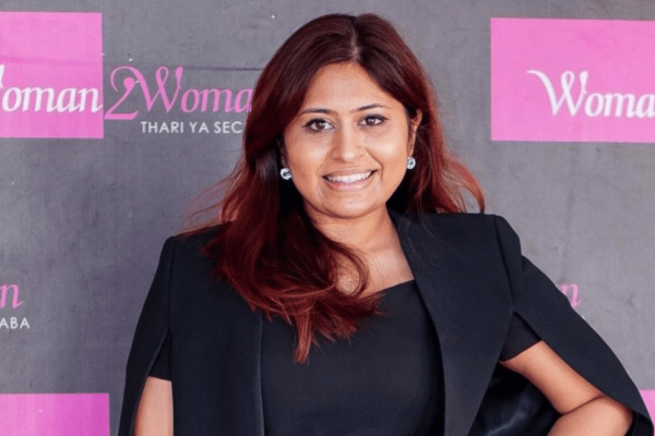 Taazima Kala-Essack, an Asian woman with chestnut coloured hair, smiles at the camera. She wears a black jacket and top. Behind her is a grey background with obscured logos in pink